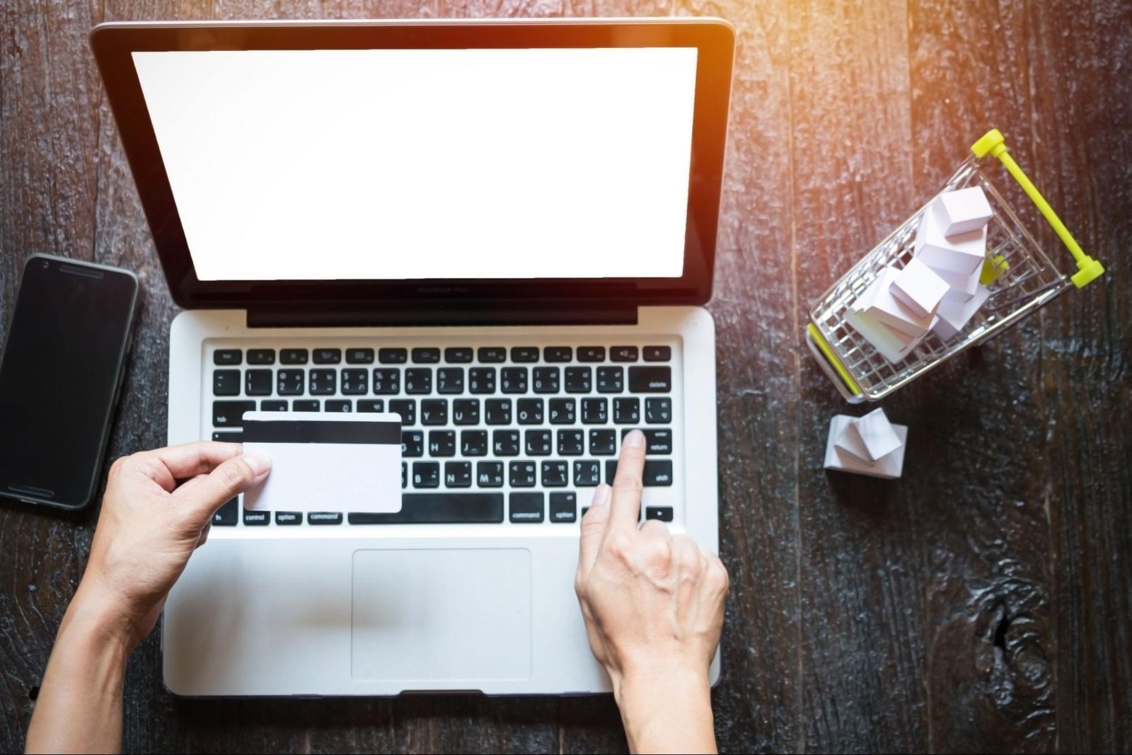 Person holding credit card while using laptop with mini shopping cart nearby, symbolizing the cost of building an ecommerce site in Pakistan.